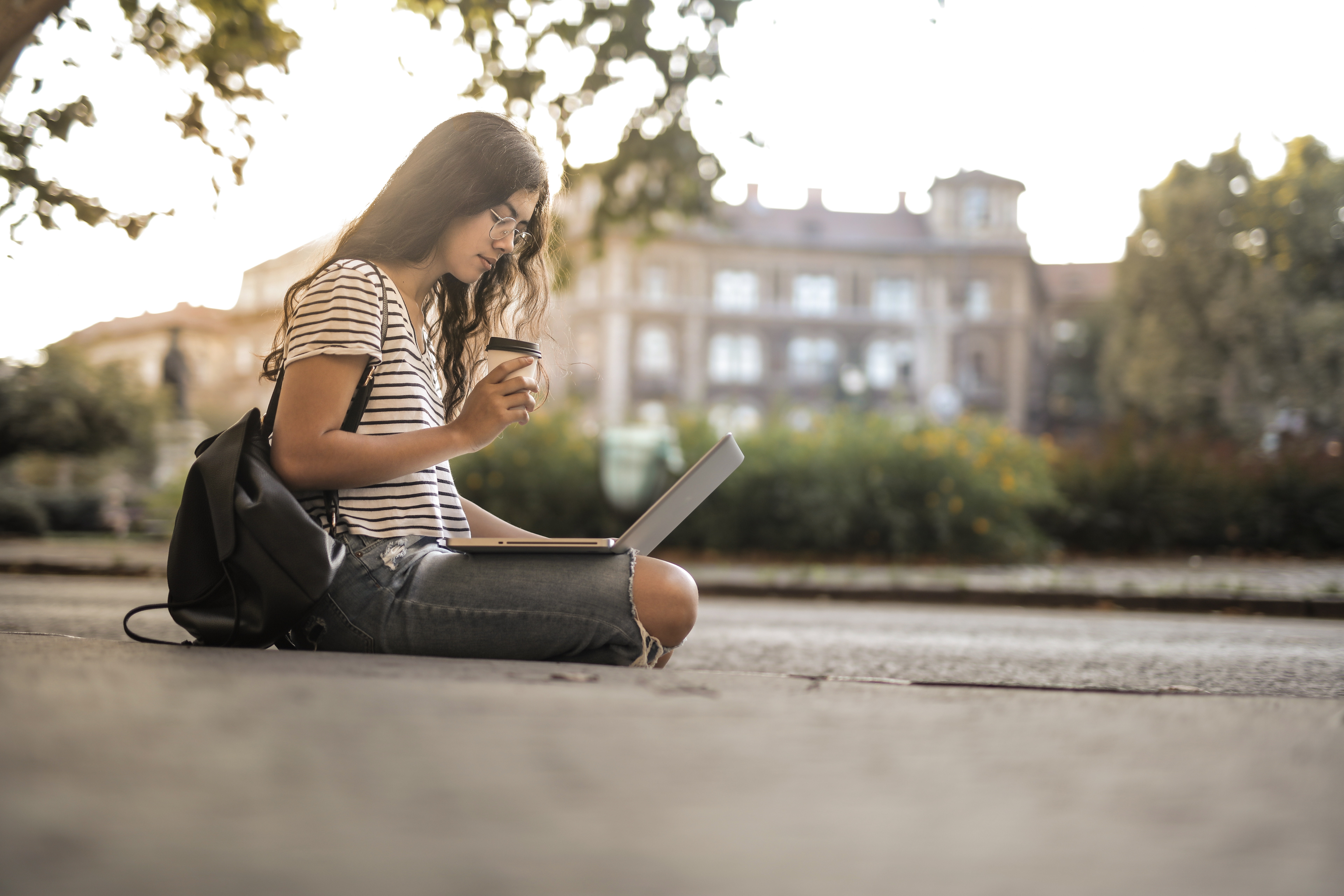 girl studying on laptop while drinking coffee and sitting on ground of college campus